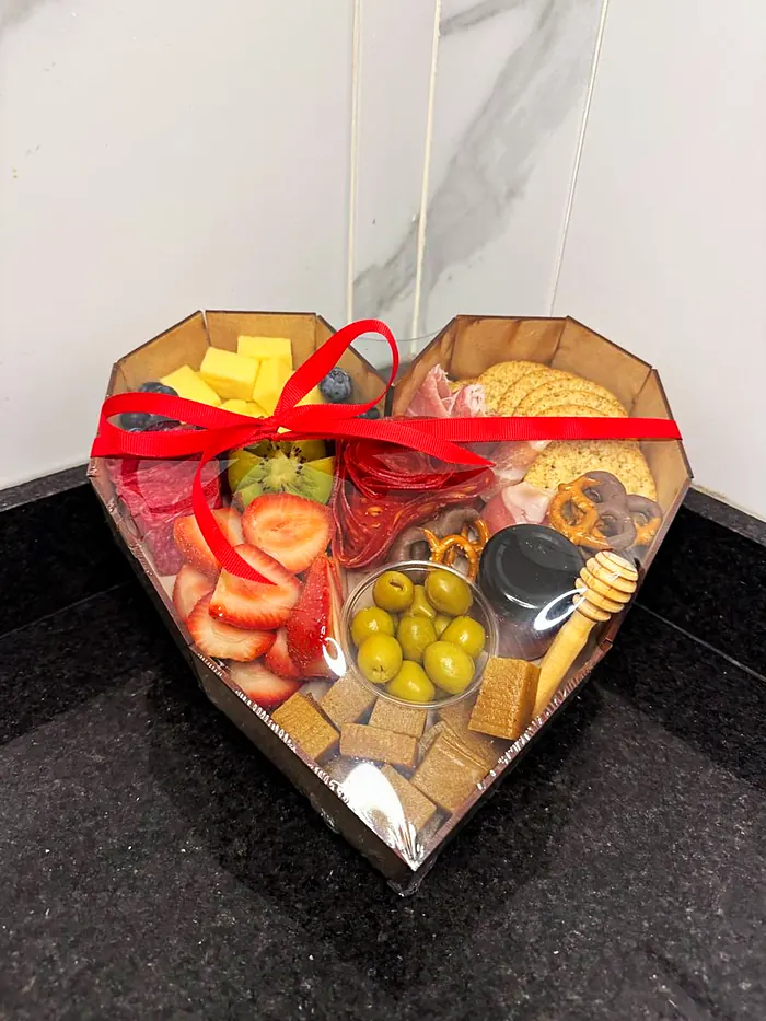 Caja en forma de corazón con fresas, quesos, carnes frías, aceitunas, pretzels y miel, con moño rojo