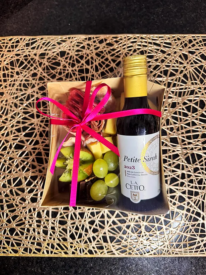 Caja individual con carnes frías, quesos, kiwi, uvas y chocolate, junto a una botella pequeña de vino tinto