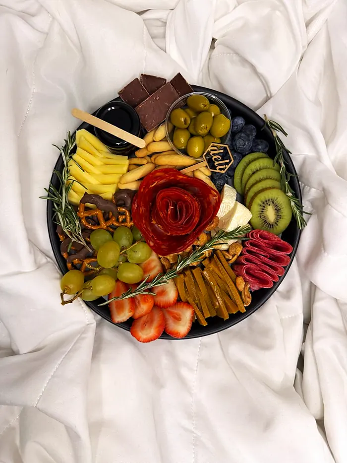 Charola redonda negra con rosa de salami, quesos, uvas, fresas, kiwi, aceitunas, chocolate y romero