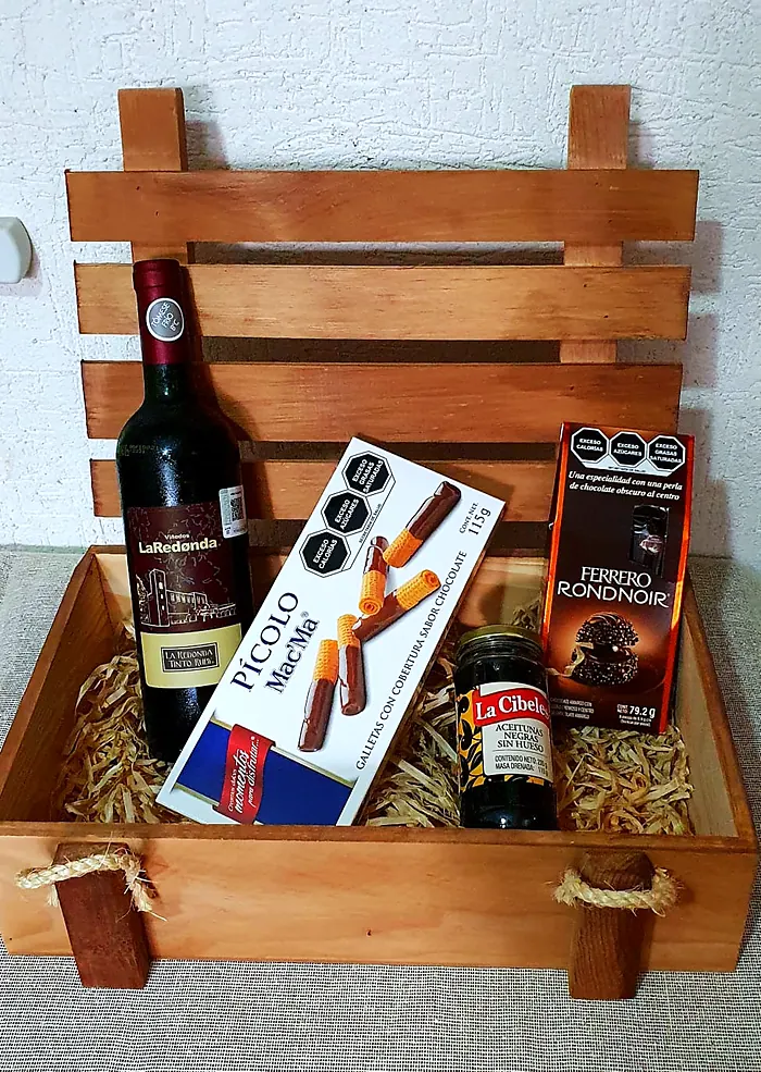 Huacal de madera con vino tinto, galletas, chocolates y aceitunas sobre paja