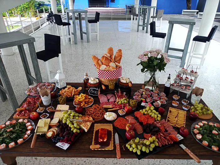 Mesa larga de madera con quesos, carnes frías, panes, frutas, uvas y postres en vasito, montada en un evento al aire libre