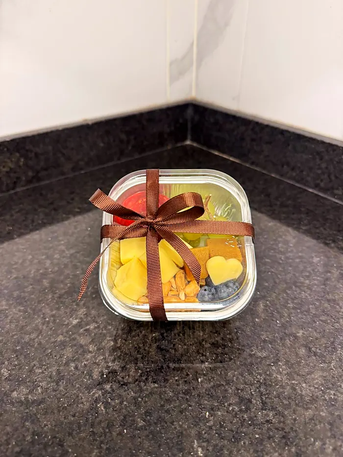 Recipiente pequeño con quesos, uvas, fresa, ate, nueces y moras, atado con moño café