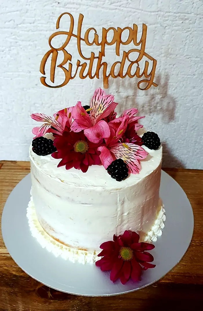 Pastel blanco semidesnudo con flores rosas, moras y un letrero de madera que dice Happy Birthday