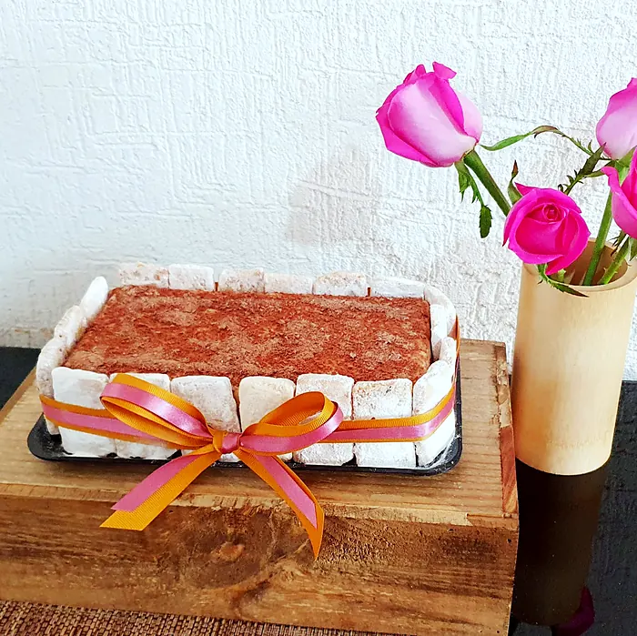 Postre rectangular rodeado de soletas con chocolate rallado, atado con listón rosa y dorado