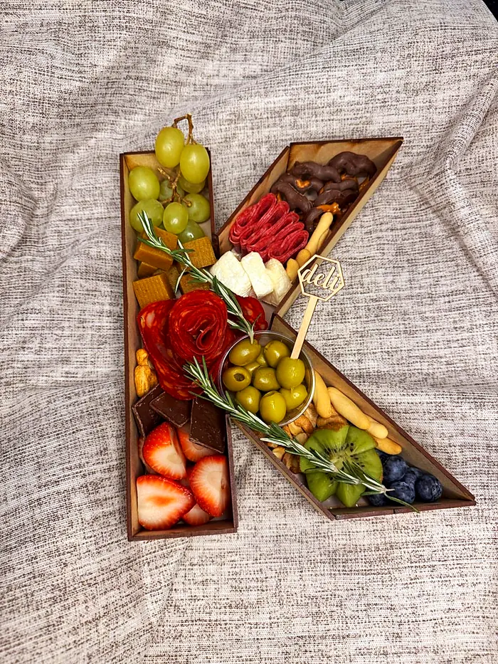 Tabla de madera con forma de letra K llena de uvas, quesos, salami, aceitunas, fresas, kiwi y moras